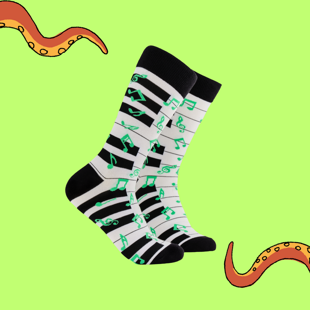 A pair of socks depicting a piano keyboard and green music notes. Black and white piano legs, black cuff, heel and toe.