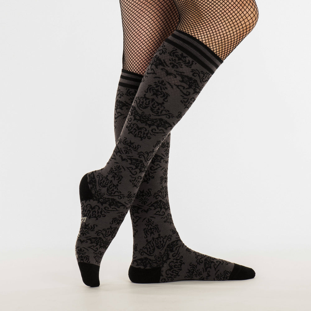 
                      
                        A model wearing a pair of knee high socks with a bat and damask pattern. Grey legs, black, heel, toe and cuff. 
                      
                    