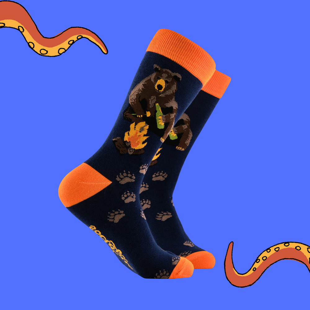 A pair of socks depicting a bear sitting by a campfire. Dark blue legs, orange cuff, heel and toe. 