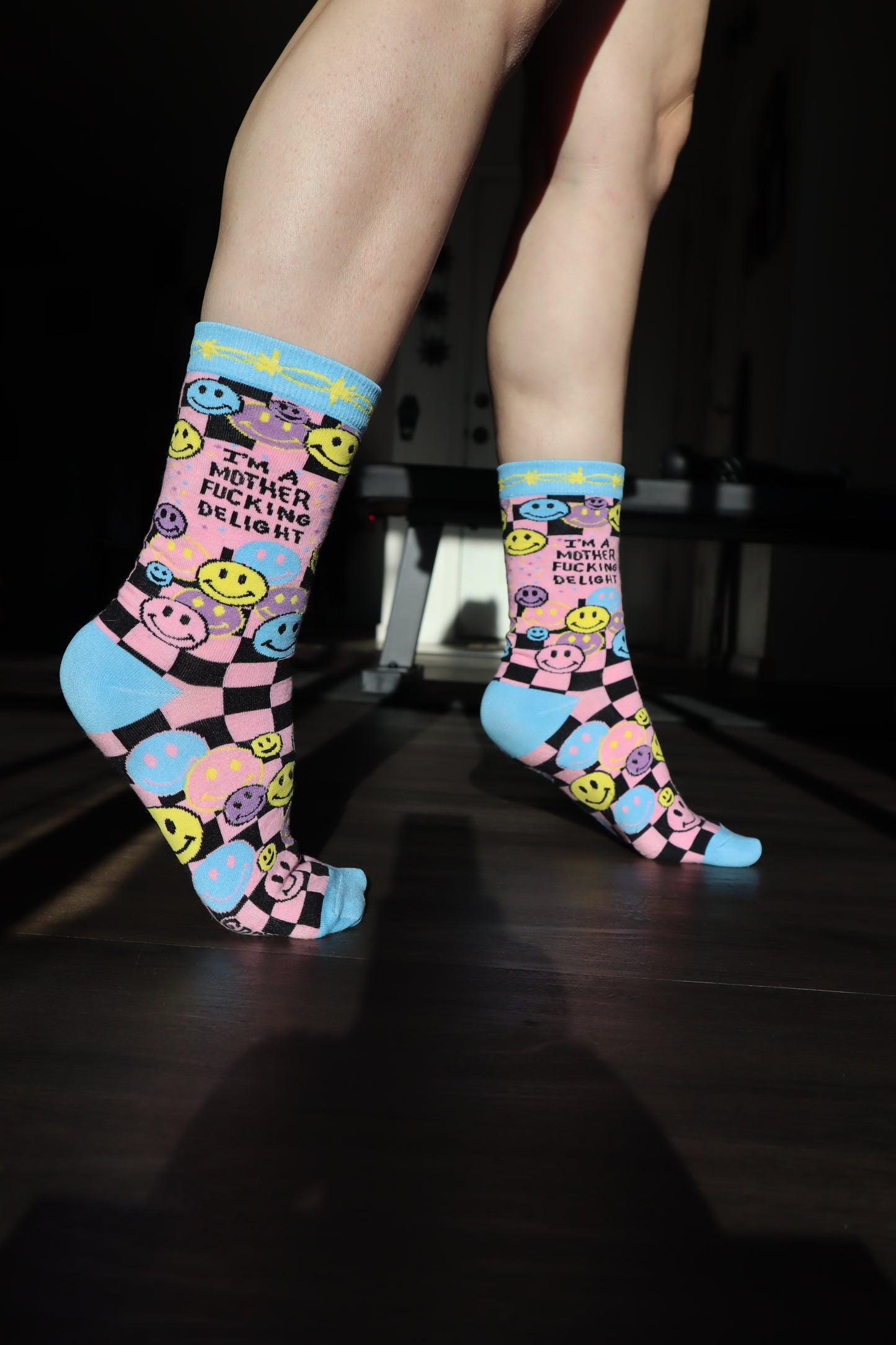 
                  
                    Model wearing a pair of socks with a bright pink and blue smiley face pattern and a bold sweary slogan. Blue toes, heel and cuff.
                  
                