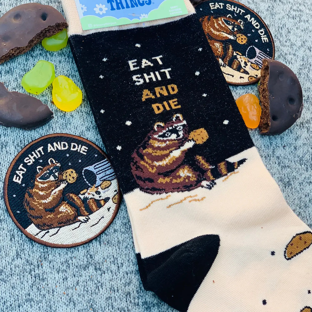 
                  
                    Eat S**t and Die Socks. A pair of socks with a bold slogan and racoon eating out of the bin detail. Black toe, cuff and heel.
                  
                