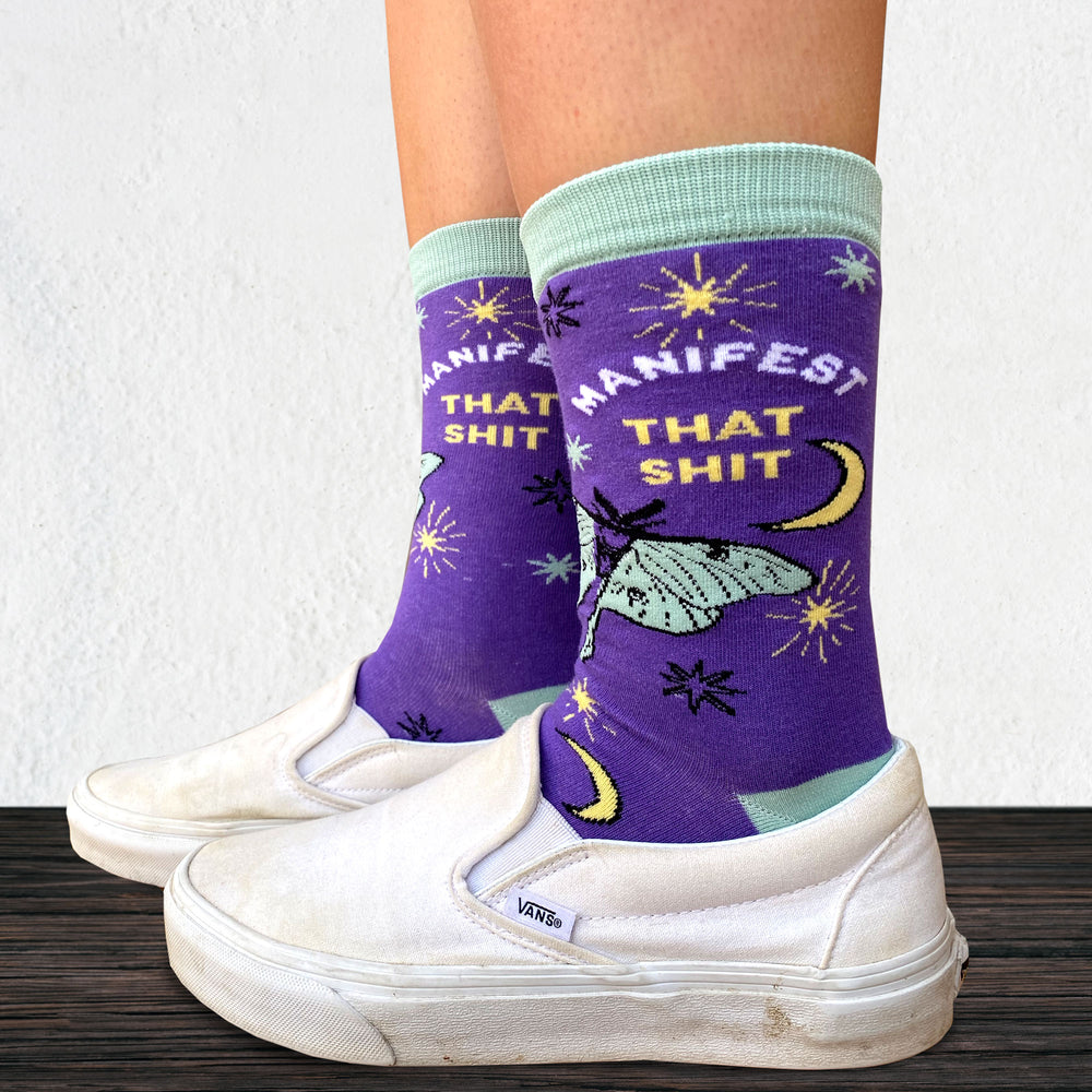 
                  
                    Model wearing a pair of socks with the slogan Manifest That S**t and a butterfly motif. Purple ankles, turquoise toe, heel and cuff.
                  
                