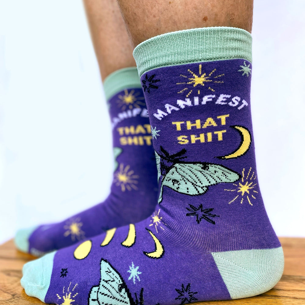 
                  
                    Model wearing a pair of socks with the slogan Manifest That S**t and a butterfly motif. Purple ankles, turquoise toe, heel and cuff.
                  
                